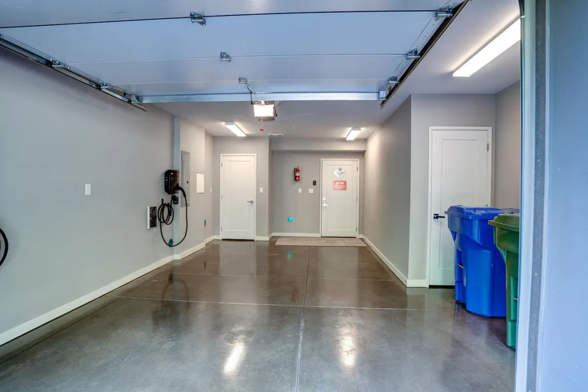 A spacious garage interior, featuring a prominent wall-mounted EV charger, a modern garage door, interior doors leading to other parts of the home, and colorful recycling bins. The setup is organized and functional.