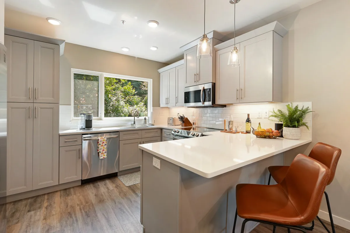 A modern kitchen with a central island, bar stools, cabinets, and expansive window views. Morning sunlight floods the room, while people cook together, suggesting lively entertaining and special occasions. The kitchen seamlessly connects to the living space, emphasizing the open-concept design.