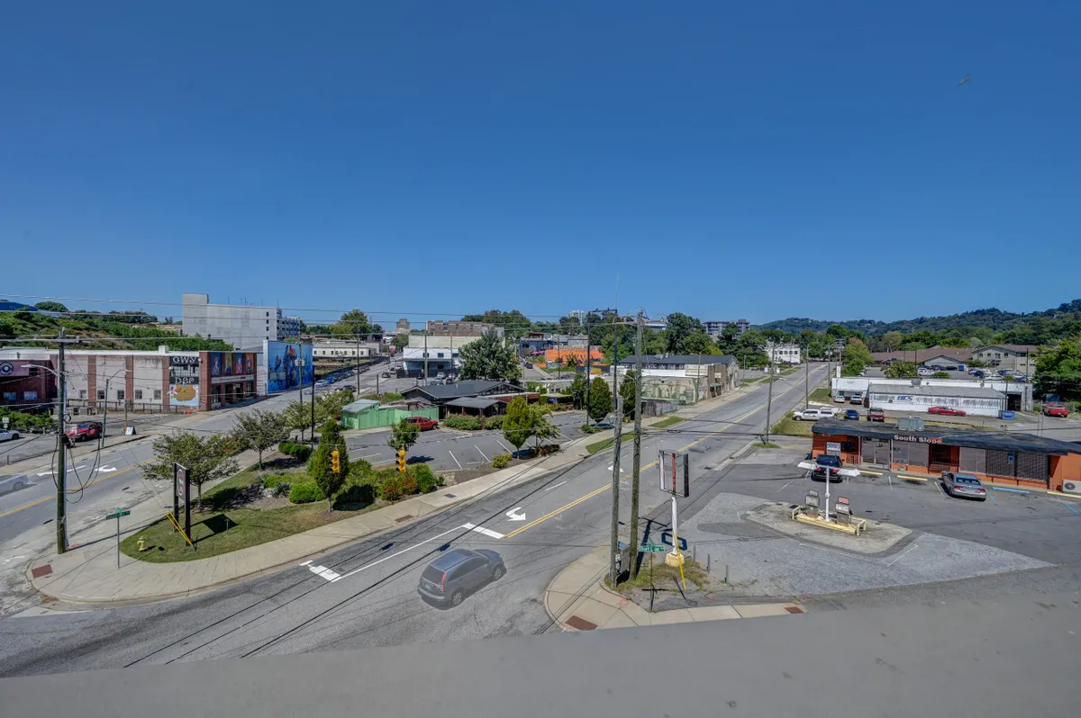 A lively city street intersection in Asheville's South Slope, featuring modern buildings, outdoor seating, greenery, and visible street art. The area is bustling with activity, hinting at the vibrant brewery and arts scene.