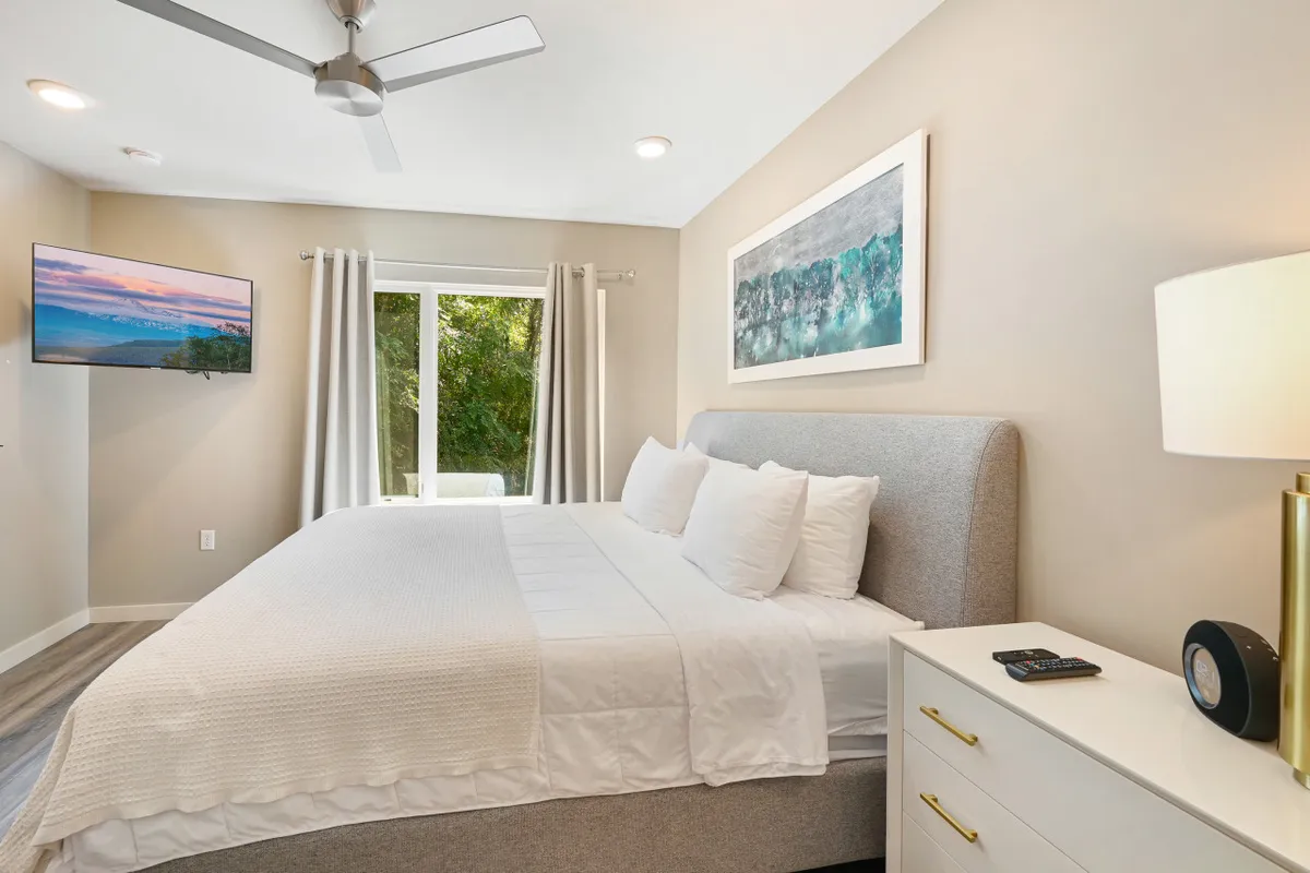 A serene king bedroom featuring a large bed with crisp white linens, artwork above the headboard, a wall-mounted TV, and curtains framing a sunlit window. The room exudes a peaceful, restful atmosphere ideal for relaxation.