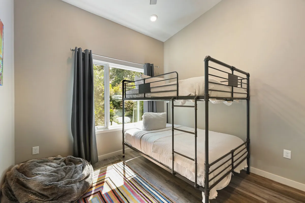 A playful bunk room featuring a sturdy bunk bed, large window streaming in natural light, a cozy bean bag chair, and a colorful rug. The layout invites both relaxation and play—ideal for children or young guests.