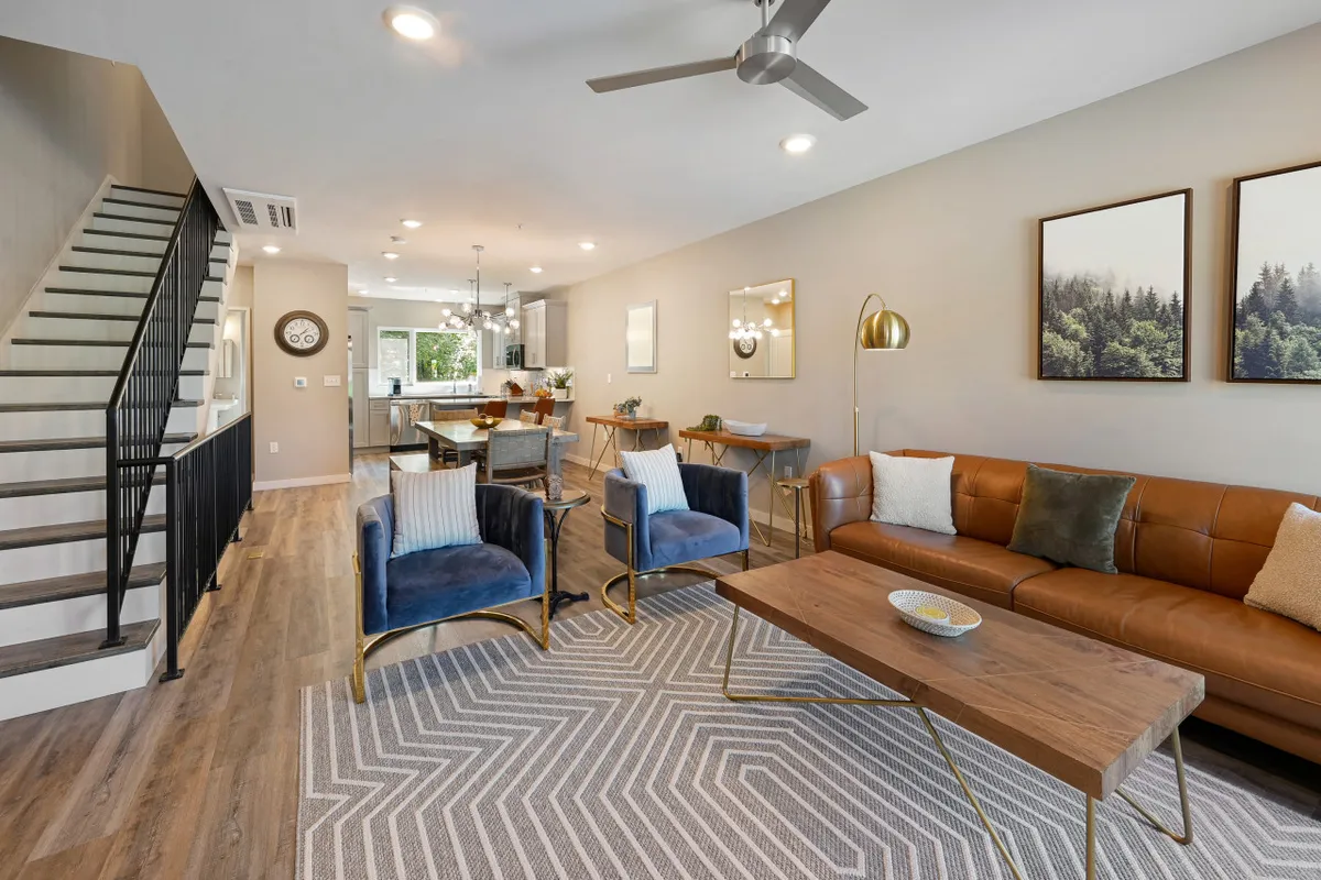 A welcoming living room featuring a leather sofa, blue accent chairs, a wooden coffee table, and a large staircase in the background. The space is designed for social gatherings and family time, exuding an inviting and cozy atmosphere ideal for relaxation or entertaining guests.