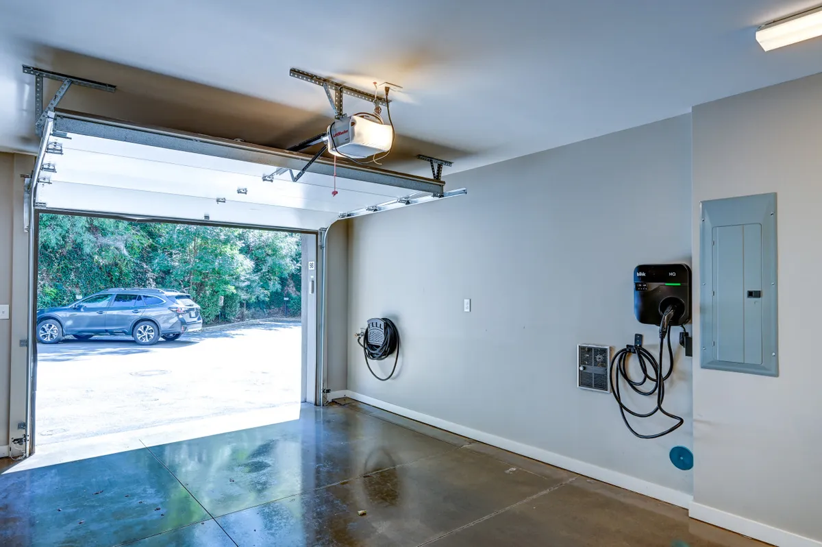A bright, modern garage featuring a clean concrete floor, visible electric vehicle charger mounted on the wall, and a neat, clutter-free space. The garage door is partially open, and the setup highlights convenient access and contemporary amenities.