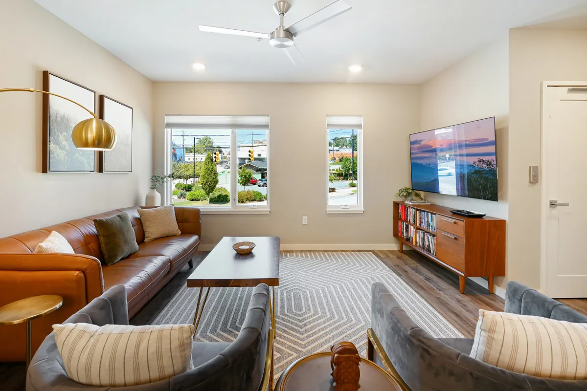 A cozy living room with a leather sofa, coffee table, TV, side chairs, and large windows letting in ample natural light. The windows offer a glimpse of the surroundings, connecting the interior space with the outside city atmosphere.