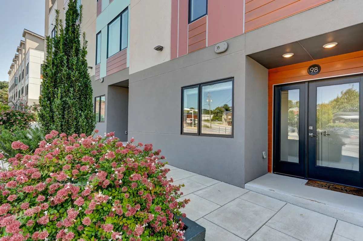 A view of the townhome's modern facade with vibrant flower bushes and a prominent front door labeled ‘98’. The image features the building set against an urban backdrop, conveying a sense of arrival and the excitement of city living. Colorful landscaping and clean lines hint at both sophistication and warmth.