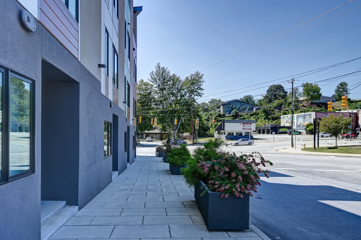 A contemporary building facade with stylish planters and a welcoming sidewalk, inviting residents and visitors to socialize or explore nearby shops. The sunny atmosphere emphasizes a sense of urban vibrancy.